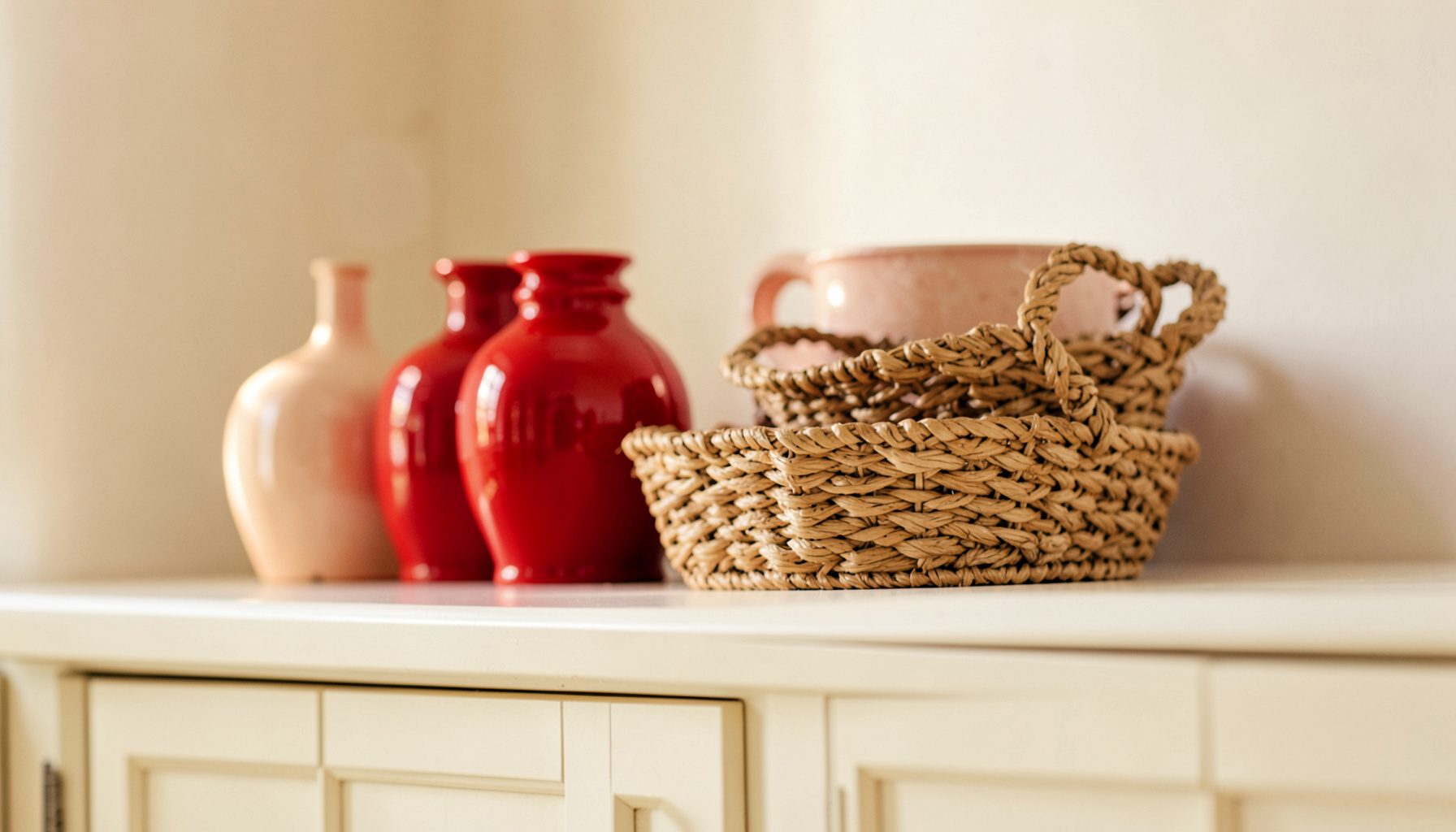 A warm, soft-lit photograph of decorative ceramic vases and woven baskets arranged on top of cream-painted kitchen cabinets, with pale ivory walls and gentle daylight